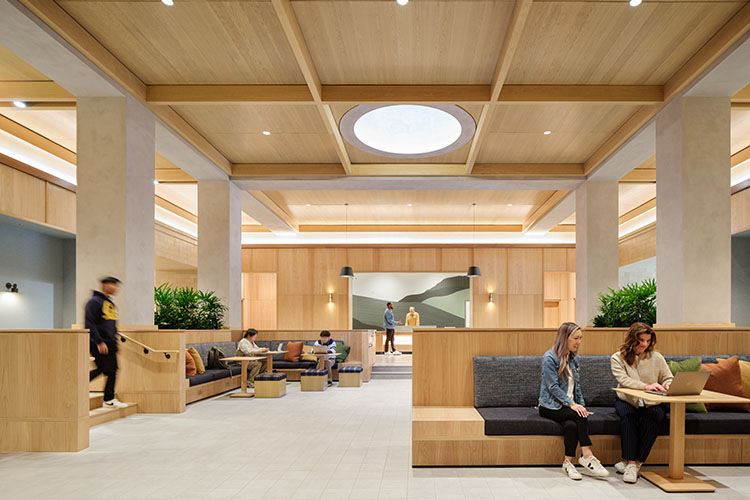 Several pairs of students sit together on sofas throughout a large indoor community space. One student is talking to a person behind the front desk at the back of the room.