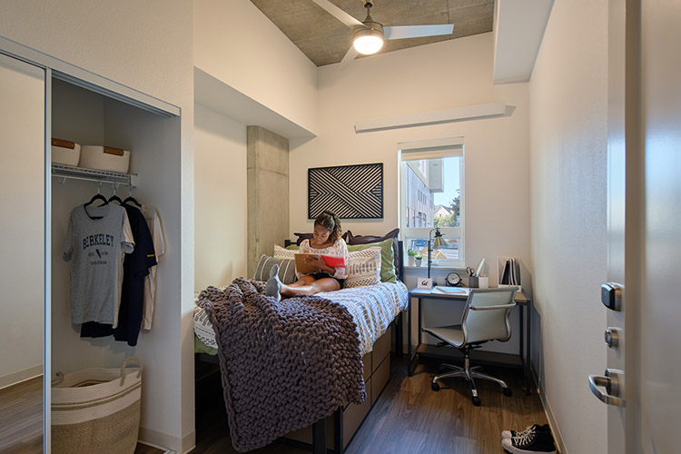 A student lies on a bed writing in a notebook. There is a a closet adjacent to the bed, as well as desk and chair in the corner of the room.