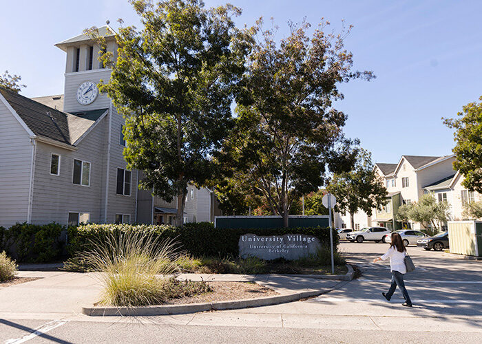 A woman walks across a parking lot towards an apartment complex with a sign titled “University Village” at the front of it.