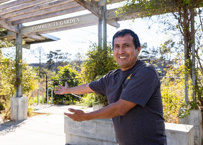 A middle-aged man outstretches his arms, pointing towards a variety of green plants in the community garden.