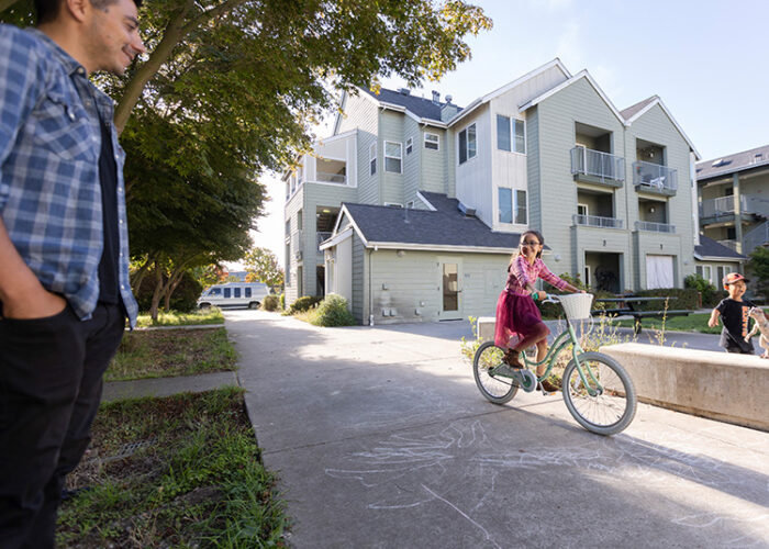 A young girl on a bike rides down the sidewalk while another young boy plays with a toy dinosaur next to her. An older male caretaker looks down at them, smiling.